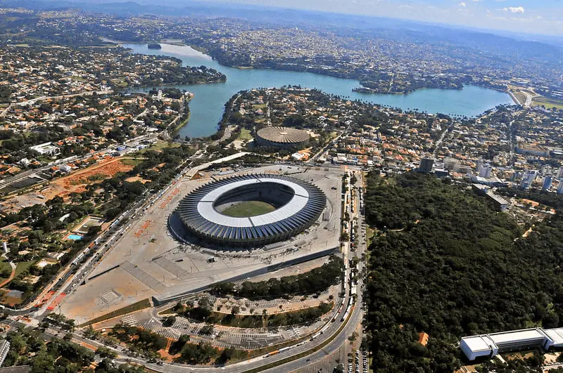 Imagem com uma visão panorâmica do Estádio Mineirão e arredores. Na imagem é possível visualizar o estádio e a esplanada, além da Lagoa da Pampulha e parte da Mata da Universidade Federal de Minas Gerais